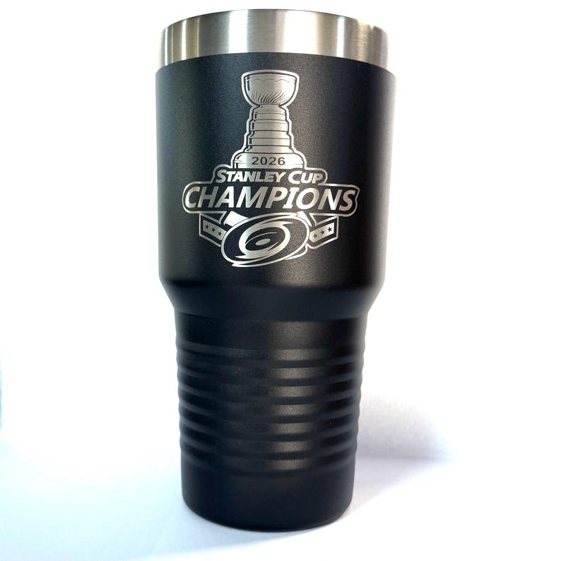 K&ouml;nnte beinhalten: Ein mattschwarzer Edelstahlbecher mit einer silbernen Lasergravur des Stanley Cup, dem Text 2026 STANLEY CUP CHAMPIONS und einem Hurrikan-Logo. Der Becher hat einen metallischen Rand und horizontale Rillen im unteren Bereich des K&ouml;rpers.