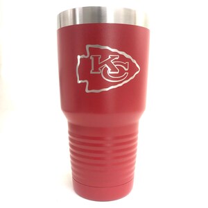 May include: A red insulated tumbler with a silver rim and a silver Kansas City Chiefs logo. The tumbler is cylindrical with horizontal ridges near the bottom. The logo is a silver outline of the team's arrowhead and the letters "KC".