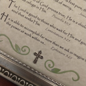 Puede incluir: Una impresión enmarcada con un marco plateado y un dije de cruz. La impresión presenta un fondo beige con acentos florales verdes y un texto inspirador de la Biblia. El texto dice: "El Señor es bueno para los que le esperan y bueno para el alma que le busca. Lamentaciones 3:25"