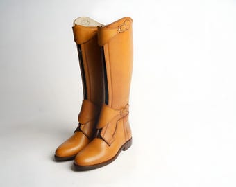 Könnte beinhalten: Ein Paar hohe, hellbraune Lederreitstiefel. Die Stiefel haben ein klassisches Design mit Schnallen, Reißverschlüssen und einer glatten Oberfläche. Die Stiefel sind etwa 40 cm hoch.