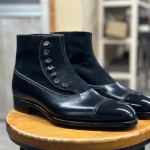 May include: Black leather ankle boots with a cap toe design. The boots have a suede panel with button closures. They are displayed on a wooden stool. The boots have a classic, formal style, suitable for smart occasions.