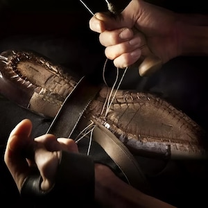 May include: A brown leather shoe sole being hand-stitched with a thick white thread. The sole features visible stitching channels and a textured surface. A wooden handle tool is used to pull the thread through the leather, securing the components.