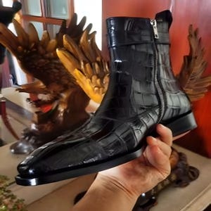 May include: A black leather boot with a crocodile texture. The boot has a side zipper and a small strap near the top. The boot is held by a hand. The boot has a square toe and a low heel.