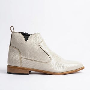 May include: An off-white ankle boot featuring a textured snakeskin pattern across the upper, a side zipper closure, a dark elastic panel near the collar, a pull tab at the heel, and a flat, light brown wooden heel with a dark rubber base.
