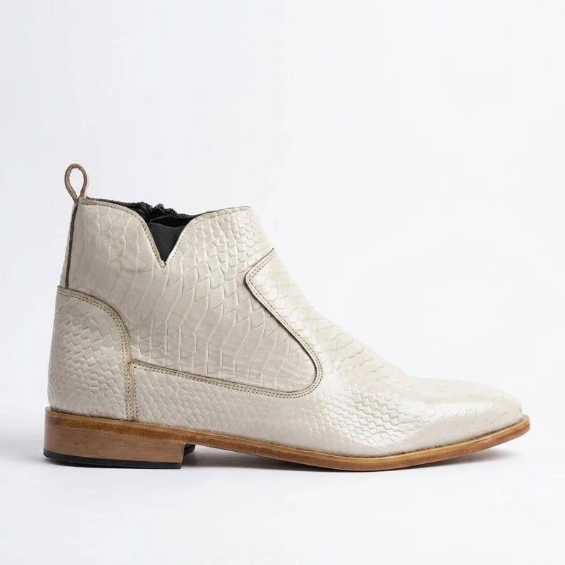 May include: An off-white ankle boot featuring a textured snakeskin pattern across the upper, a side zipper closure, a dark elastic panel near the collar, a pull tab at the heel, and a flat, light brown wooden heel with a dark rubber base.