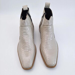 May include: A pair of off-white ankle boots featuring a textured snakeskin pattern, a pointed toe, a side zipper closure, and a pull tab at the heel. The boots have a smooth finish, a dark interior lining, and a light brown sole edge.
