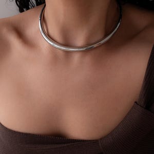 May include: A polished silver choker necklace. The necklace is a simple, curved design that sits close to the neck. The jewelry is displayed on a person wearing a brown top.
