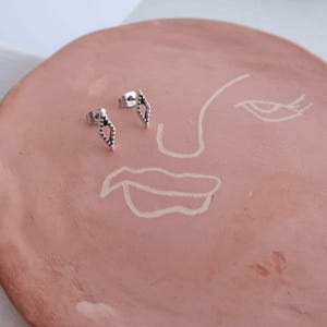 Può includere: Un paio di orecchini a lobo in argento con un design geometrico. Gli orecchini sono su un piatto color terracotta con un disegno a linea bianca di un viso.