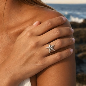 Puede incluir: Un anillo de plata con forma de estrella de mar. El anillo presenta un diseño detallado de estrella de mar, con cinco puntas y una superficie texturizada. El anillo se lleva en un dedo, con el océano al fondo. Es una joya.
