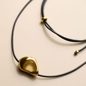 Op de afbeelding: Een goudkleurige hangerketting met een gladde, organische vorm. De hanger hangt aan een zwart koord, met een kleine gouden kraal accent. De ketting heeft een verstelbaar ontwerp.