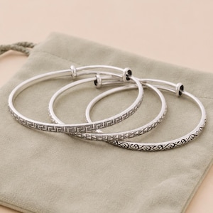 Op de afbeelding: Een set van drie uitbreidbare armbanden in zilverkleur. Elke armband heeft een eigen gegraveerd patroon: één met een Grieks sleutelontwerp, één met een getextureerd piramidemotief en één met een decoratief krulpatroon.