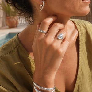 May include: A set of silver-toned jewellery including a curved earring, a thin textured band ring, a wider ring with a central oval accent, and a double-layered cuff bracelet. The jewellery features a polished metallic finish and smooth, organic shapes.