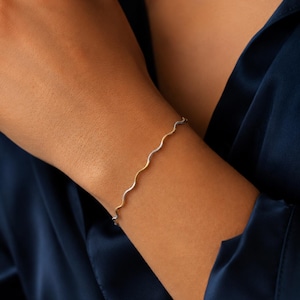 May include: A thin, wavy bangle bracelet featuring an alternating two-tone design of polished silver-coloured metal and gold-coloured metal. The bracelet has a continuous, fluid, serpentine shape that wraps around the wrist in a simple, delicate style.