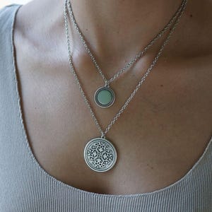 May include: Two silver necklaces with round pendants. The top pendant is a small, light green circle. The bottom pendant is a larger, silver circle with an ornate, swirling design. The necklaces are layered.