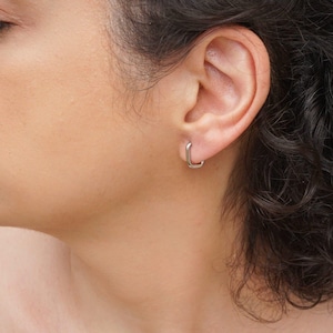 Puede incluir: Primer plano de un pendiente cuadrado plateado. El pendiente es de diseño pequeño y minimalista, y se lleva en la oreja. El pendiente tiene una forma geométrica simple.