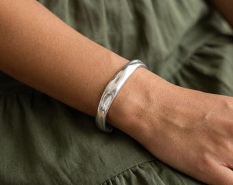 May include: A silver-toned cuff bracelet with a smooth, polished finish and an irregular, organic shape. The bracelet features a wide, open-ended band design that wraps around the wrist, showcasing a reflective surface and a solid, continuous metal construction.