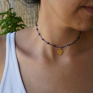 Puede incluir: Un collar de oro con un colgante redondo cuelga de un cordón azul con pequeñas cuentas azules. El collar lo lleva una persona con una camiseta blanca de tirantes.