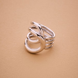 Può includere: Un anello color argento con un design astratto unico. L'anello presenta più fasce curve che si avvolgono attorno al dito, creando un effetto a spirale. Il metallo lucido riflette la luce, evidenziando la forma scultorea dell'anello. L'anello è posto su uno sfondo neutro.