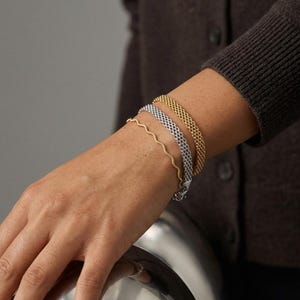 Pode incluir: Um conjunto de três pulseiras usadas juntas no pulso. O conjunto inclui uma pulseira fina de fio dourado ondulado, uma pulseira de corrente de malha prateada e uma pulseira de corrente de malha dourada mais larga, com acabamento metálico.