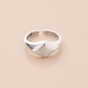 Può includere: Un anello color argento con un design geometrico. L'anello presenta un elemento centrale a forma di diamante e una fascia aperta. Il numero "17" è inciso sulla fascia. L'anello è posto su uno sfondo neutro.