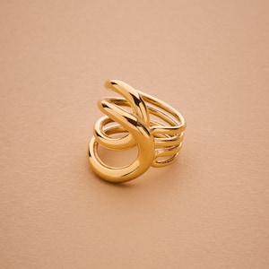 Op de afbeelding: Een goudkleurige ring met een uniek, wervelend ontwerp. De ring heeft meerdere banden die buigen en overlappen, waardoor een opvallende, sculpturale uitstraling ontstaat. De ring is gemaakt van een glanzend, reflecterend metaal.