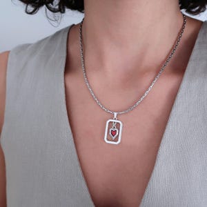 Op de afbeelding: Zilverkleurige ketting met een rechthoekige hanger met een rood hartontwerp. De hanger hangt aan een fijne ketting. De ketting wordt gedragen op een effen gekleurde top.