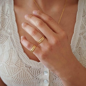 May include: A hand wearing three gold rings. Two rings are on the index finger and one on the pinky finger. The rings have a band of small, clear stones. The person is wearing a white, open-knit sweater and a gold necklace.