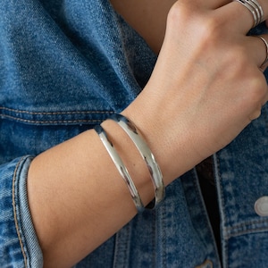 May include: Two silver bangle bracelets stacked on a person's wrist. The bracelets are simple and sleek, with a smooth, polished finish.