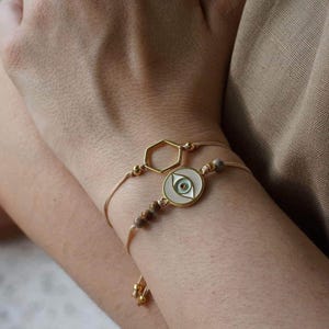 May include: Two gold bracelets on a wrist. One bracelet has a gold hexagon charm and the other has a round charm with a blue and green evil eye design.