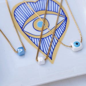 May include: Four gold chain necklaces with different blue and white ceramic charms. The charms are shaped like squares and feature an evil eye design.