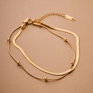 Op de afbeelding: Een goudkleurige, gelaagde armband met een slangenkettingontwerp. De armband heeft een delicate ketting met kleine, ronde kralen en een tweede, bredere ketting. De sluiting en verlengketting zijn ook goudkleurig.