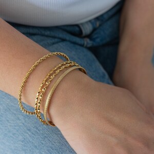 Può includere: Quattro braccialetti d'oro impilati sul polso di una persona. I braccialetti sono di larghezze diverse e hanno diverse texture. I braccialetti sono un articolo di gioielleria popolare.