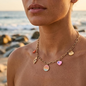 Könnte beinhalten: Goldfarbene Kette mit mehreren Anhängern. Zu den Anhängern gehören ein korallenfarbenes Stück, eine Muschel, ein Anhänger mit bunten Streifen, ein rosa Herz mit Augendesign und eine Muschel. Die Kette wird vor einem verschwommenen Strandhintergrund getragen.