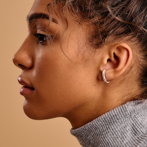 Könnte beinhalten: Seitenprofil einer Person mit grauem Rollkragenpullover und silbernem Creolen-Ohrring. Der Ohrring hat eine kleine, strukturierte Halbmondform. Der Hintergrund ist einfarbig beige.