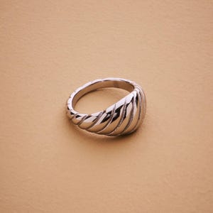 Könnte beinhalten: Ein silberfarbener Ring mit geripptem Design. Der Ring hat eine abgerundete Form und eine polierte Oberfläche, die das Licht reflektiert. Der Ring ist vor einem neutralen Hintergrund platziert.