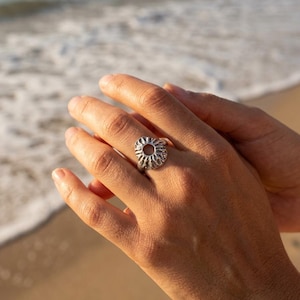 Puede incluir: Un anillo plateado con un diseño circular y texturizado que se asemeja a una criatura marina. El anillo se lleva en una mano, con un fondo borroso de playa y océano.