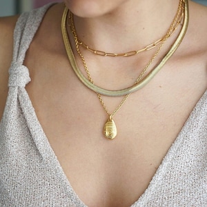 Może przedstawiać: Złoty naszyjnik w kilku warstwach z zawieszką o szczegółowym wzorze. Naszyjnik nosi osoba w jasnobrązowym topie.