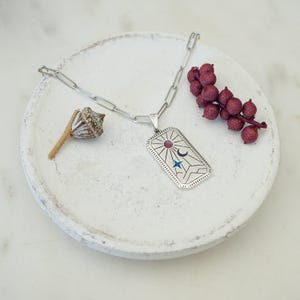 Puede incluir: Un collar de plata con un colgante rectangular que presenta un diseño de sol, luna y estrella. El colgante tiene un aspecto texturizado y grabado.