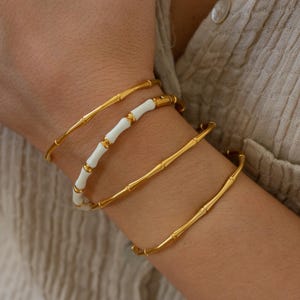 Op de afbeelding: Een set van drie gouden armbanden in bamboestijl. Een armband heeft witte emaille accenten.