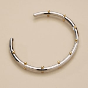 May include: A silver cuff bracelet with small, evenly spaced gold beads. The bracelet has a smooth, polished finish and a simple, open design. Jewellery.