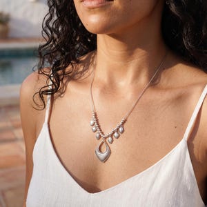 Peut inclure: Un collier argenté doté d'un pendentif central en forme de goutte avec une surface texturée, suspendu à une chaîne délicate. La chaîne est ornée de plus petites breloques en forme de goutte assorties sur les côtés, créant un design symétrique.