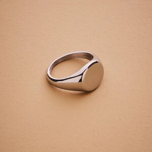 May include: A polished silver signet ring with a smooth, oval face. The ring is set against a neutral, tan background, highlighting its reflective surface. The ring's design is simple and classic, suitable for everyday wear.