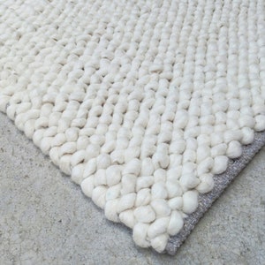 Puede incluir: Primer plano de una alfombra blanca y esponjosa con un dise&ntilde;o tejido texturizado. La alfombra tiene un borde gris claro.