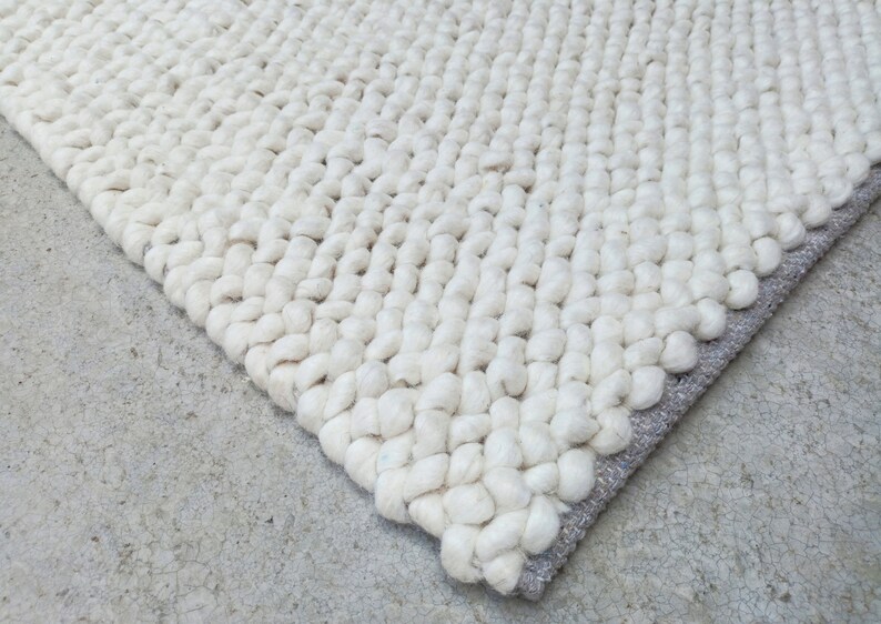 Puede incluir: Primer plano de una alfombra blanca y esponjosa con un dise&ntilde;o tejido texturizado. La alfombra tiene un borde gris claro.