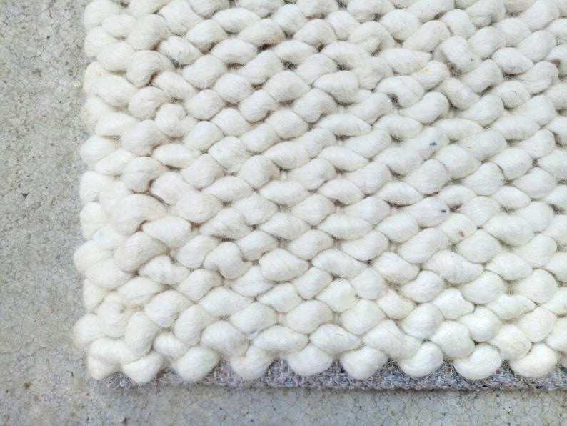 Puede incluir: Alfombra blanca tejida con un patr&oacute;n de punto grueso y texturado. La alfombra tiene un respaldo gris claro.