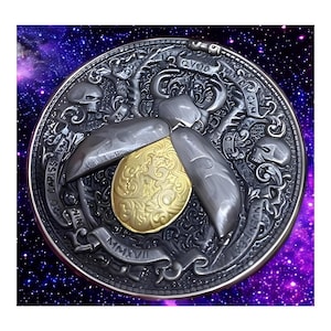 May include: A silver and gold coin with a detailed design of a beetle with wings spread. The coin has the words "QVOD ERAT IN TENEBRIS" and "MMXVII" inscribed on it.