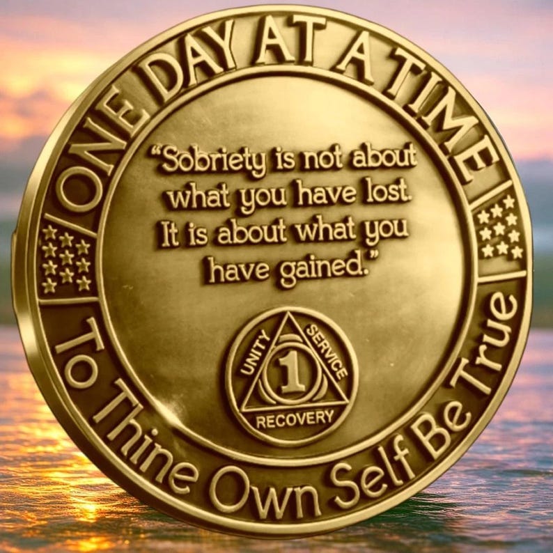 May include: A circular gold-toned sobriety medallion featuring the text "ONE DAY AT A TIME" and "To Thine Own Self Be True" along the border. The center displays the quote "Sobriety is not about what you have lost. It is about what you have gained." and a triangle symbol with the words "UNITY SERVICE RECOVERY" and the number "1".