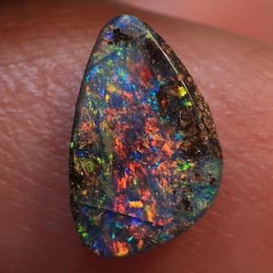 Könnte beinhalten: Ein tropfenförmiger Opal-Edelstein mit dunkler Grundfarbe und lebhaftem Farbenspiel im Inneren. Der Opal zeigt Blitze in Rot, Orange, Grün und Blau. Der Stein hat eine polierte Oberfläche und eine natürliche, organische Form.