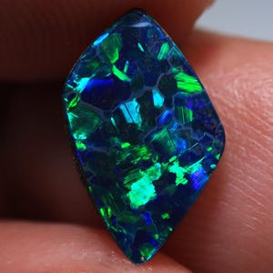 Könnte beinhalten: Nahaufnahme eines schwarzen Opal-Edelsteins in Diamantenform mit lebendigen grünen und blauen Lichtreflexen. Die inneren Muster und Farben des Edelsteins sind sichtbar. Der Opal wird zwischen Fingern gehalten.