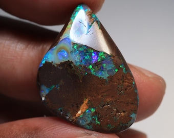 Puede incluir: Una piedra preciosa de ópalo en forma de lágrima con una matriz marrón y vibrantes destellos de azul, verde y púrpura. La superficie pulida refleja la luz, mostrando el juego de colores interno de la piedra. El ópalo se sostiene entre los dedos.